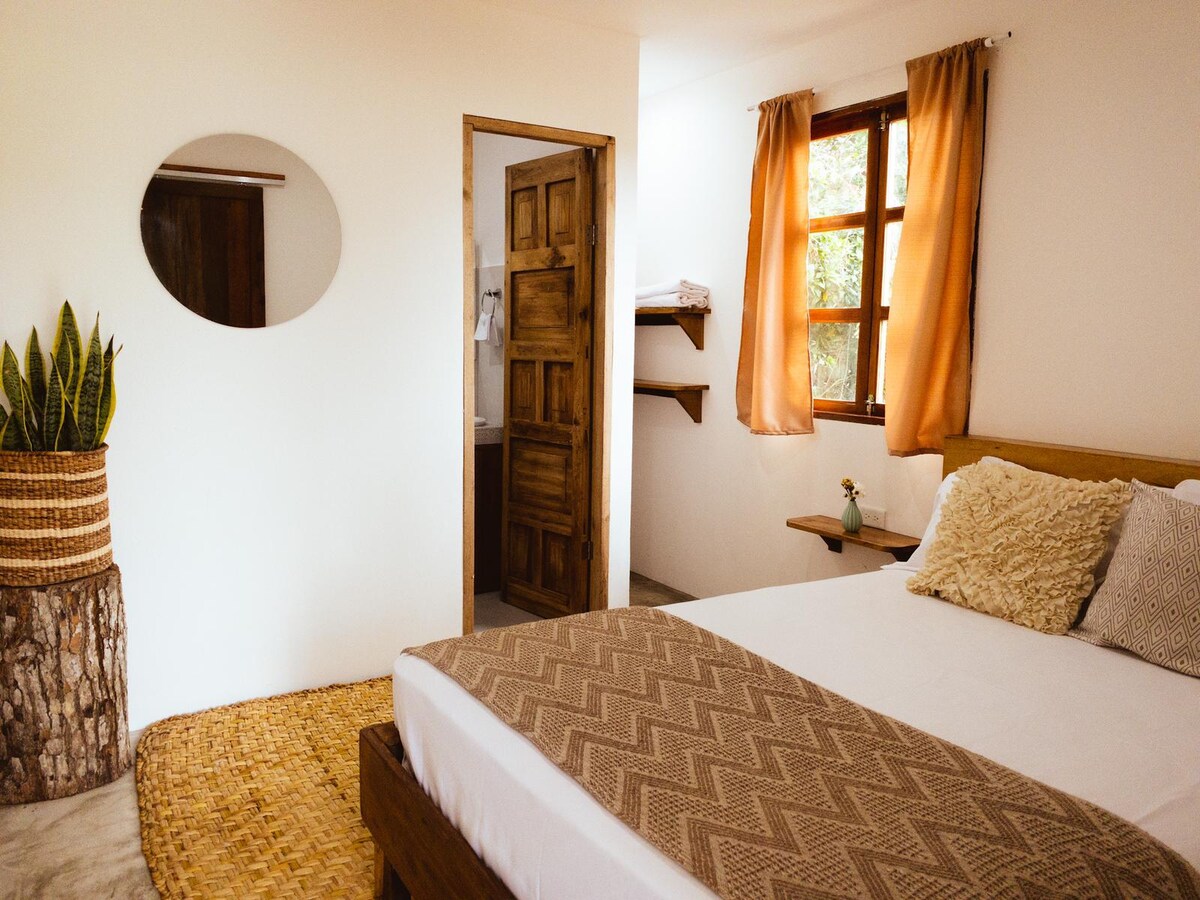 The room features a spacious bed with neutral-toned bedding and decorative pillows. A natural fiber rug lays on the floor, and a potted plant adds a touch of greenery. A wooden door and windows allow natural light to fill the space, while a simple shelf provides additional storage.