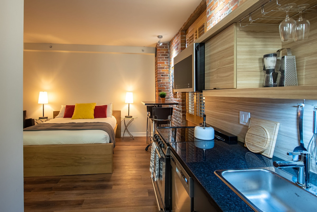 A cozy studio space features a queen bed with colorful accent pillows. A kitchenette is situated along one wall, complete with a sink and sleek countertop. Soft lighting from two lamps adds warmth, while exposed brick walls enhance the modern design.