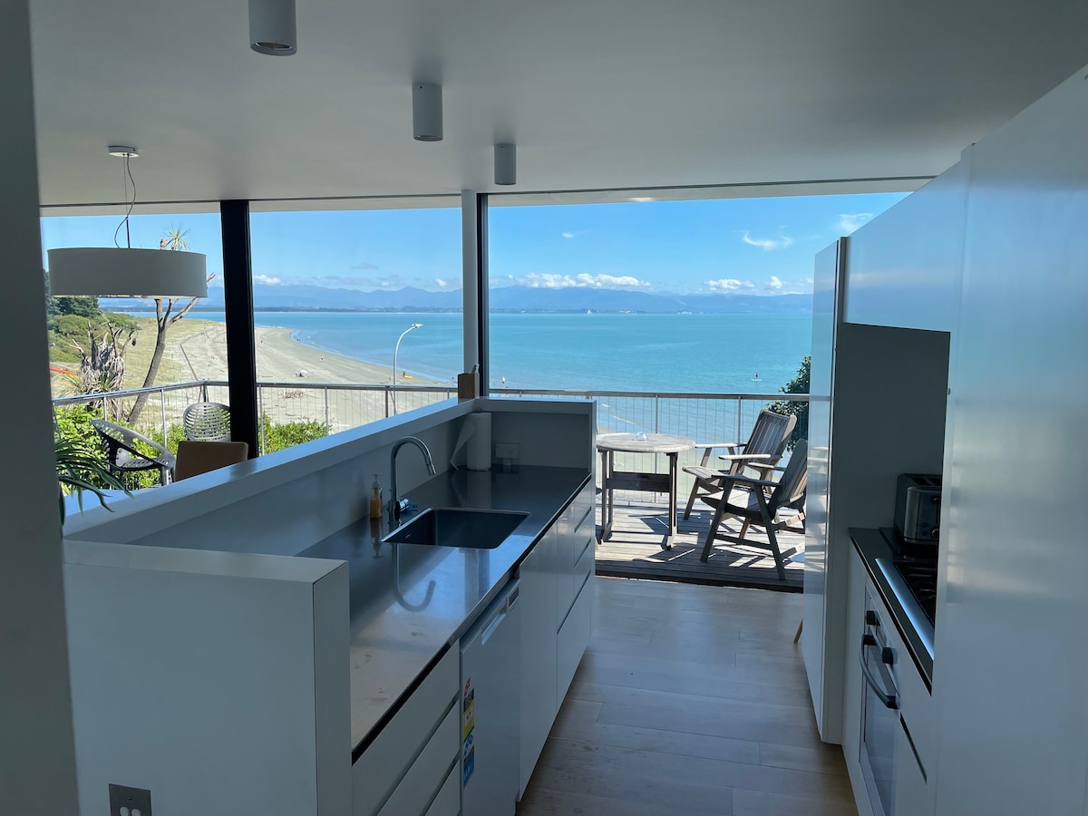 An open-plan kitchen displays modern white cabinetry and sleek appliances. Large glass windows offer a panoramic view of the beach and ocean beyond, with a dining area visible on the balcony. Natural light brightens the space, enhancing the airy atmosphere.