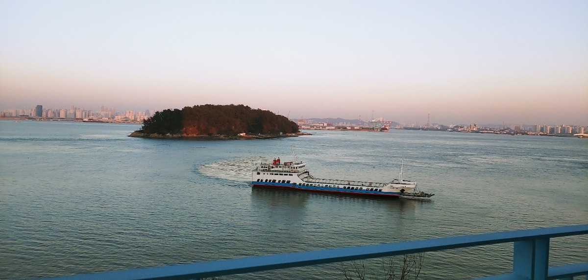 A scenic view of tranquil waters featuring a ferry navigating around a small island. The skyline of a city is visible in the background, with soft pastel hues of a sunset merging with the horizon.