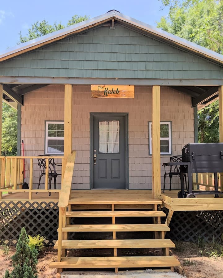 A rustic cabin exterior is presented with a light gray and green color scheme. The porch features a wooden railing and steps leading to a central door, flanked by two small tables and chairs. A grill is positioned to the right.