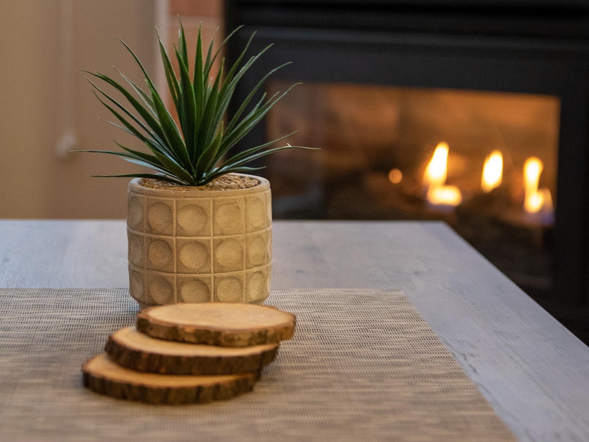 A modern table setting is displayed with a textured pot containing a green plant. Slices of wood are placed neatly on a light woven placemat, with a warm fireplace softly glowing in the background.