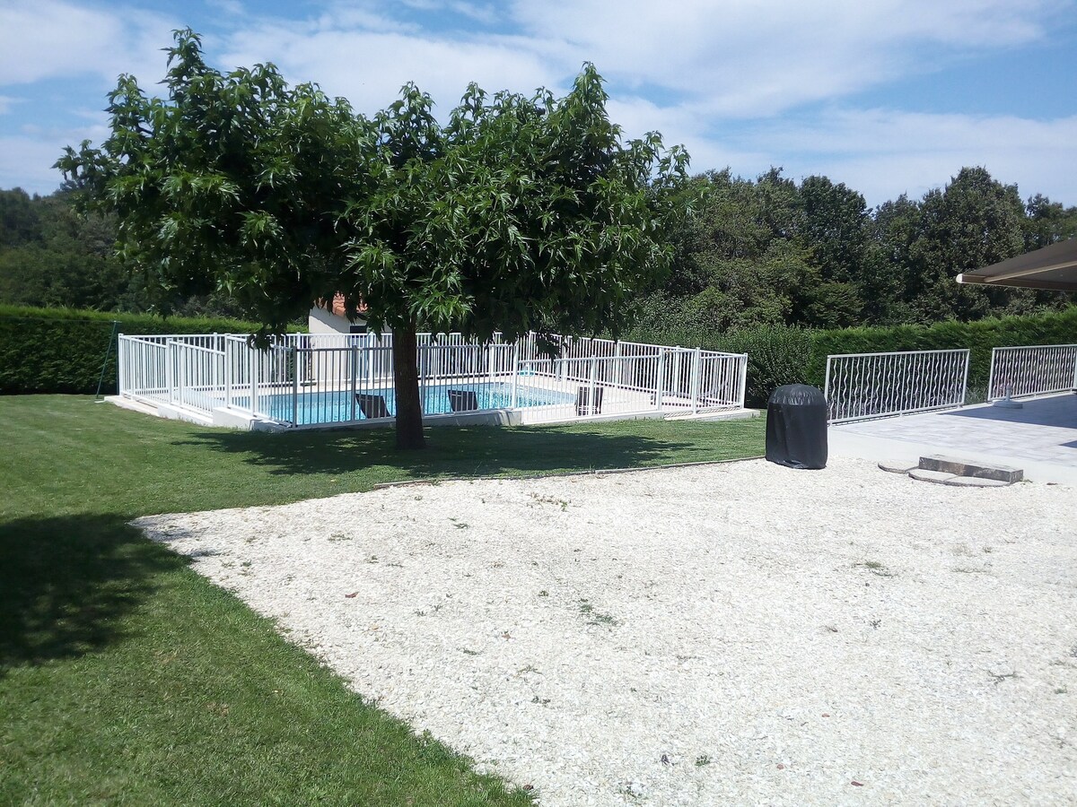 A secure swimming pool is surrounded by a white fence, set against a backdrop of lush greenery. A large tree provides shade on the grassy area, while a gravel path leads to the pool. A black barbecue grill is positioned nearby for outdoor cooking.