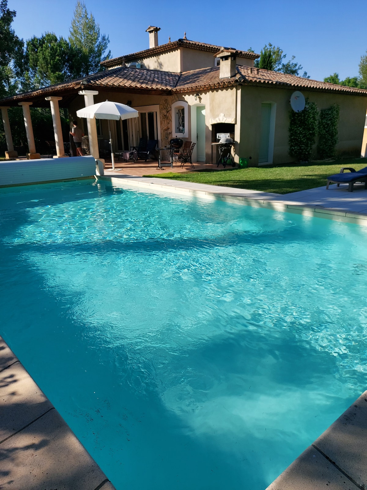 A private swimming pool glistens under the sunlight, surrounded by a tiled patio. The villa features a porch with seating and a barbecue grill, providing a relaxing outdoor space. Lush greenery is visible around the area, enhancing the tranquil environment.
