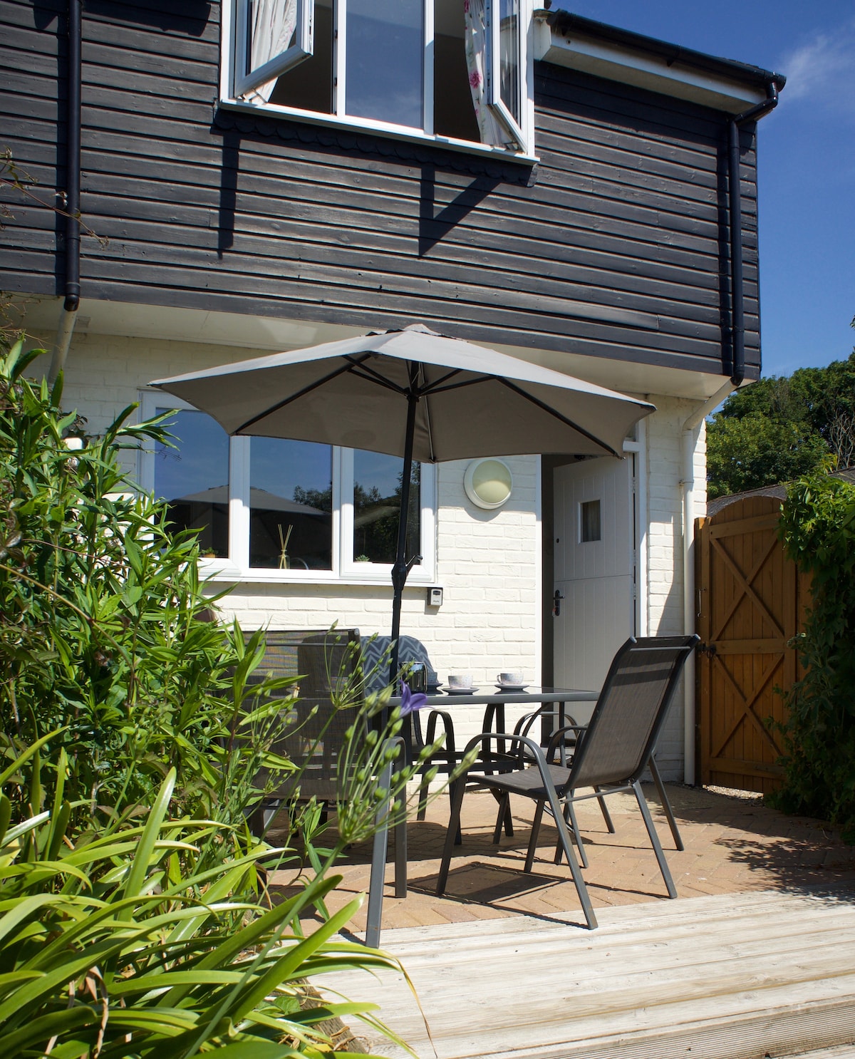 A small outdoor seating area features a table and chairs under a gray umbrella, surrounded by greenery. The entrance to the house is visible, along with a wooden gate leading to the side area. The bright sky adds to the light atmosphere.