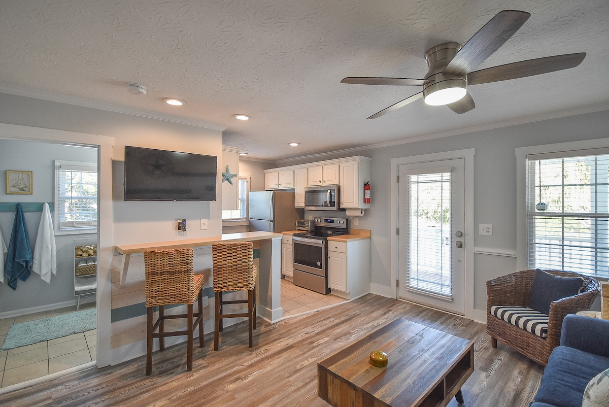 An open-concept living space features a well-equipped kitchen with stainless steel appliances and a breakfast bar with two wicker stools. A comfortable seating area is present, furnished with a cozy couch and a wooden coffee table. Natural light floods in through the glass door leading to the outdoor area.