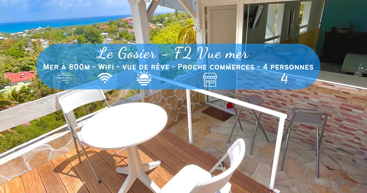 A bright terrace showcases a small outdoor dining set with two chairs and a table, framed by a railing. The stunning view features greenery and glimpses of the sea, highlighting the inviting atmosphere of the surrounding environment. Proximity to local shops is indicated.