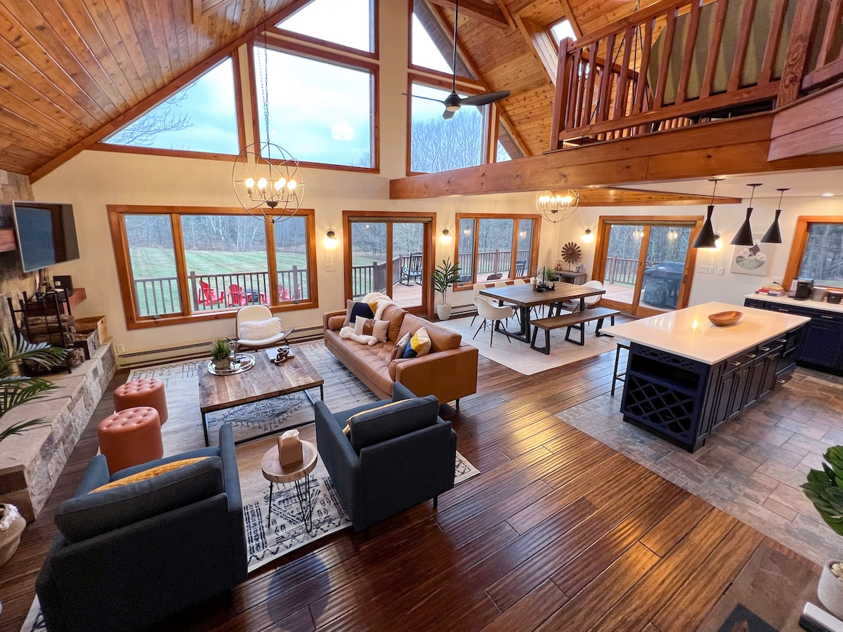 An open floor plan living area features vaulted wooden ceilings and large windows offering views of the outdoors. Comfortable seating areas include a tan couch, black chairs, and a decorative coffee table. A dining space with a large table and an island kitchen is also visible.