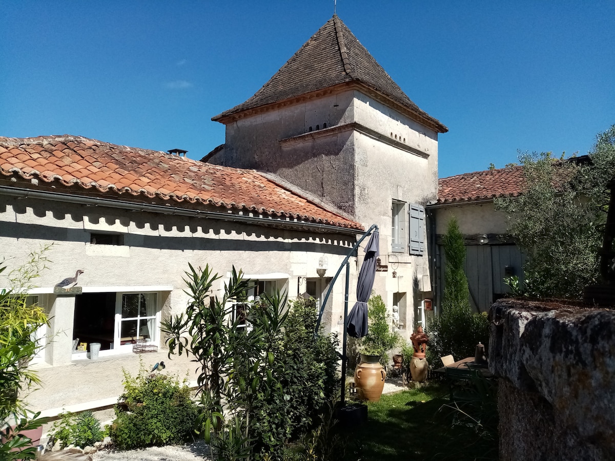 The exterior of a charming rustic building, featuring traditional stone walls and a distinctive tower with a shingled roof. Lush greenery surrounds the property, with various plants and decorative elements in the garden, all set against a clear blue sky.