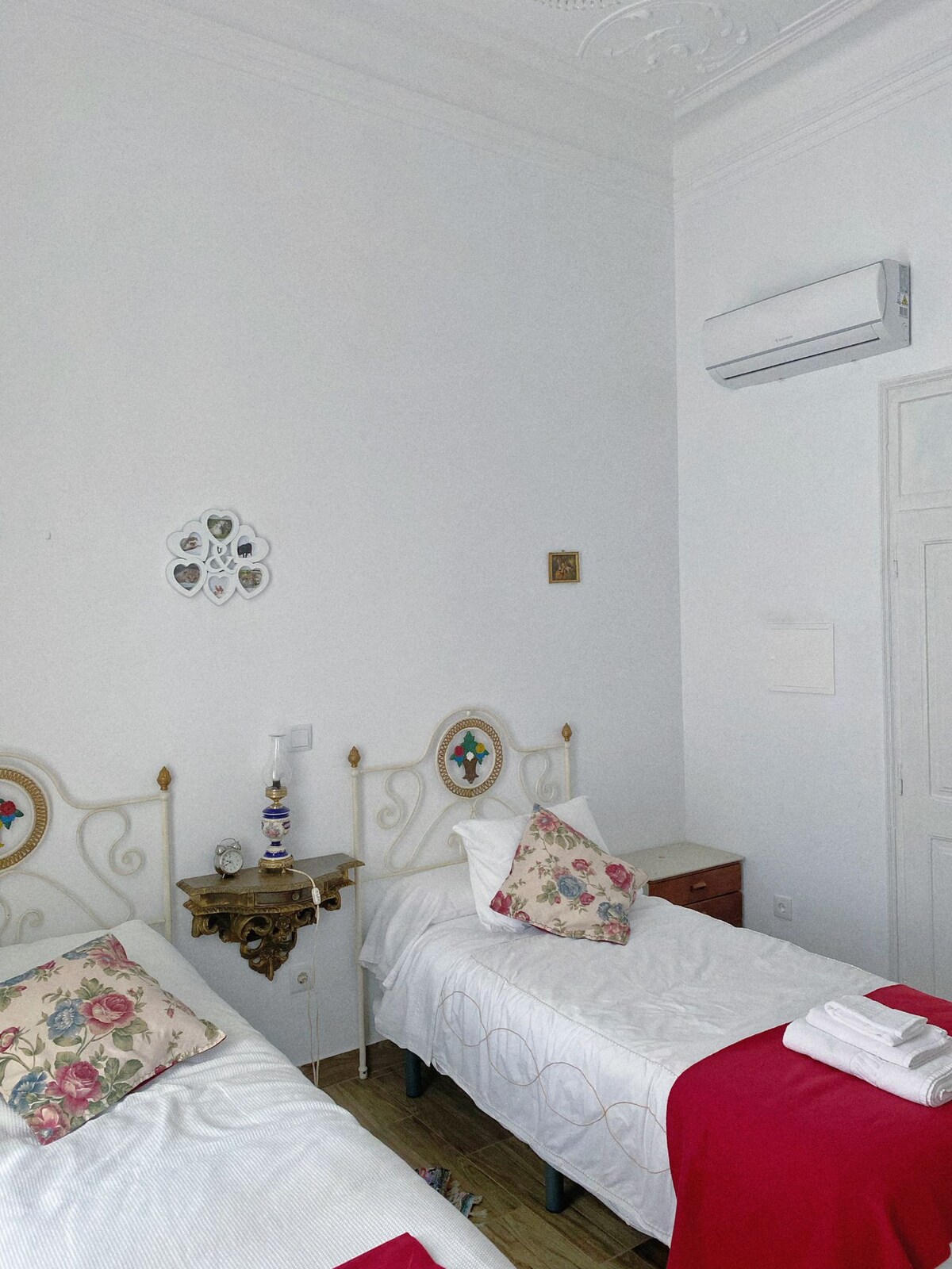 The room features two twin beds with decorative headboards, adorned with floral-patterned pillows. A small side table is placed between the beds, and a wall-mounted air conditioner provides climate control. Light walls enhance the room's spacious feel.