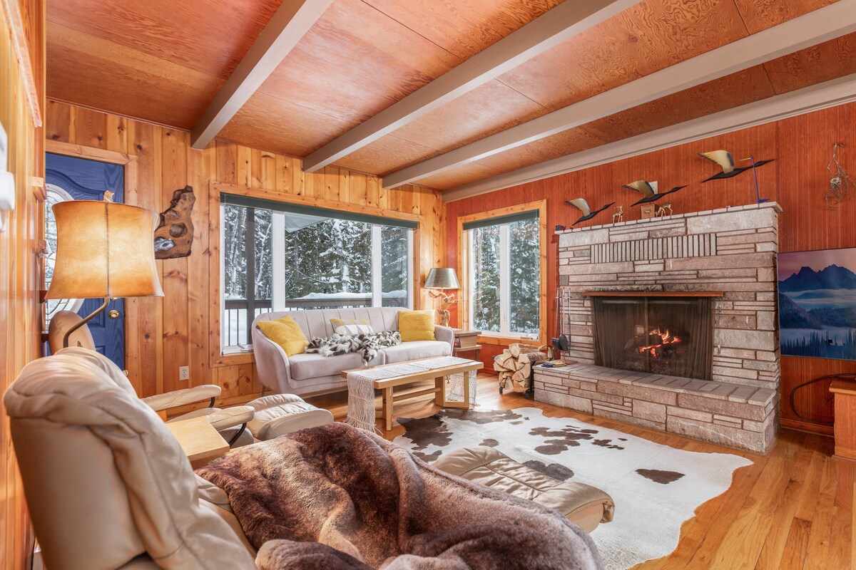 A warm living area features a stone fireplace, providing a focal point against the wooden wall. Comfortable seating includes a light-colored sofa and armchair, while a cozy throw lies on a plush chair. Large windows frame views of the snowy outdoors.