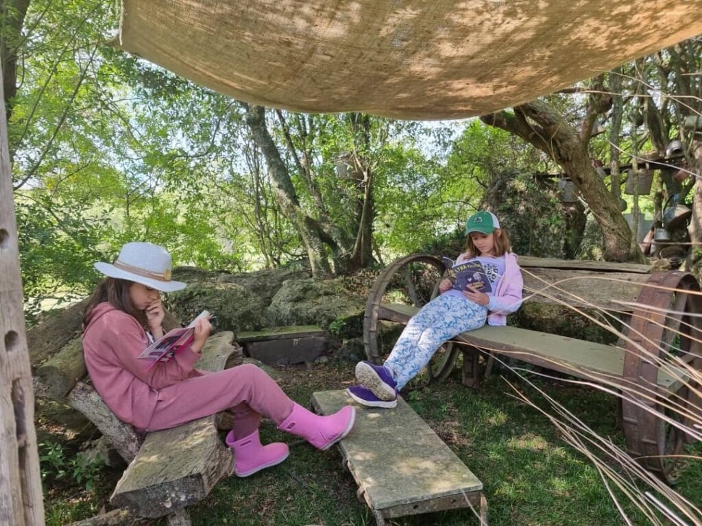 A shaded outdoor area is visible, featuring two wooden benches positioned under a canopy. Two children are sitting on the benches, each engaged in reading a book. Surrounding trees provide a natural backdrop, enhancing the serene atmosphere of the space.