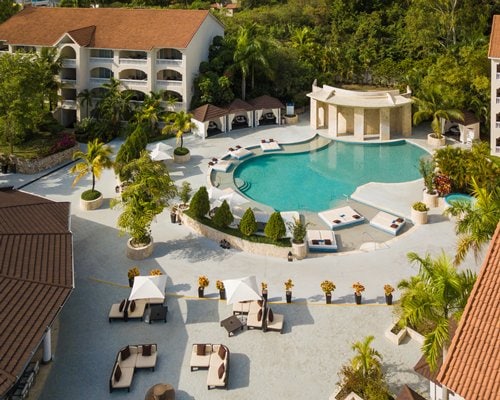 An aerial view of the resort features a spacious pool surrounded by shaded cabanas and lush landscaping. Sun loungers are placed around the pool area, enhancing relaxation options. The buildings are set against a backdrop of greenery, creating a serene atmosphere.