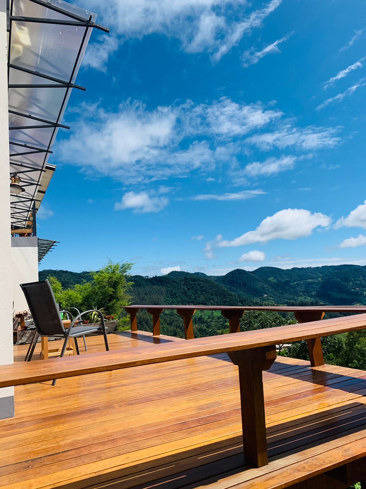 A wooden deck extends from the house, offering scenic views of the surrounding mountains under a bright blue sky. A chair is positioned on the deck, inviting relaxation while enjoying the landscape.