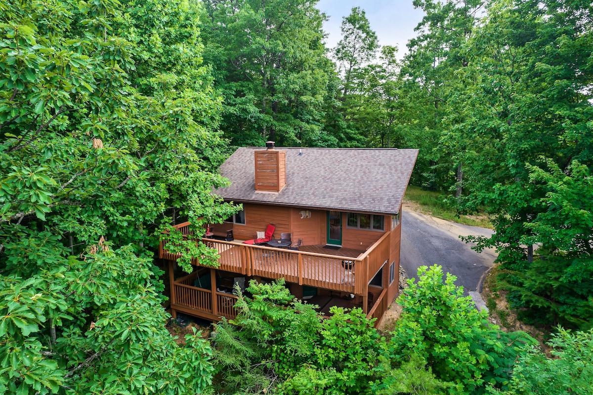 The cabin is nestled among lush greenery, featuring a spacious wraparound deck with several outdoor chairs. A tree-lined road is visible nearby, enhancing the secluded atmosphere in the Smoky Mountains community.