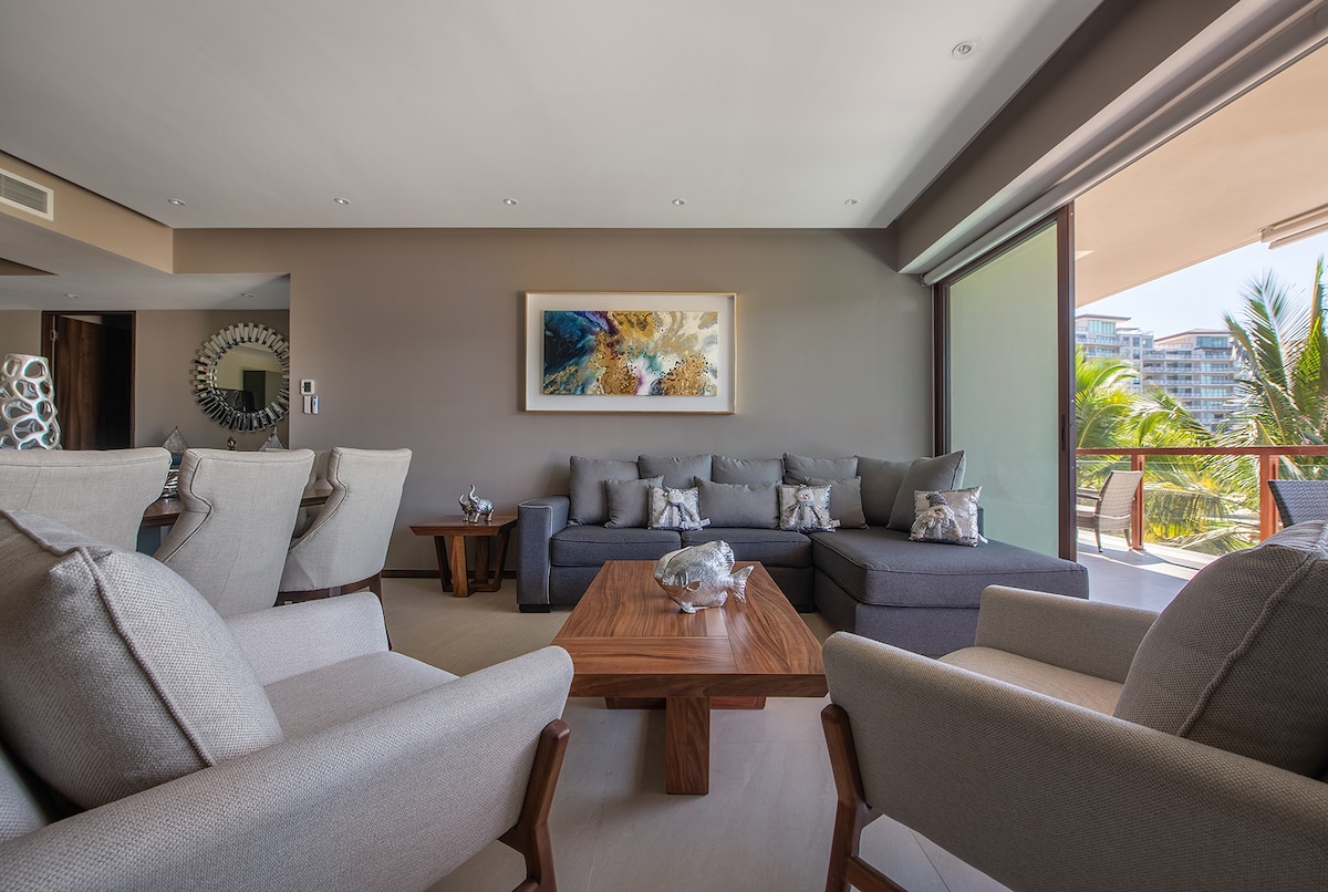 A spacious living area is furnished with a comfortable sectional sofa and accent chairs, paired with a wooden coffee table. Large glass doors invite natural light and provide access to a balcony with views of palm trees. A colorful artwork is displayed on the wall.