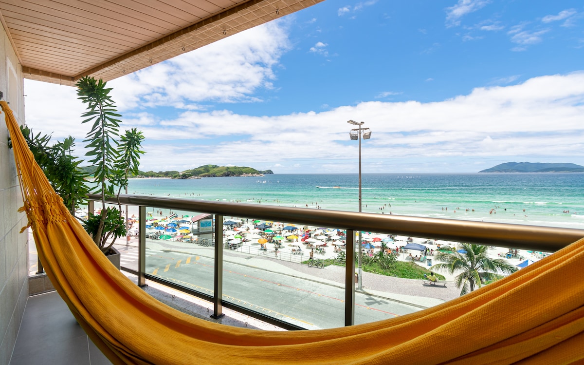 A vibrant yellow hammock is suspended on a balcony, offering a view of the ocean and beach. The scene features clear blue skies, gentle waves, and colorful beach umbrellas dotting the sandy shore, creating an inviting outdoor space for relaxation.