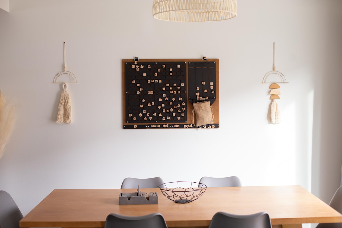 A light-filled dining area features a wooden table accompanied by gray chairs. A decorative wall board displays various textured elements, while two hanging accents add a touch of warmth to the room. A simple wire bowl sits centrally on the table.