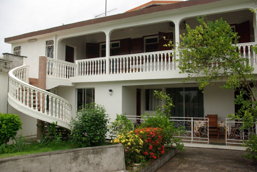 The exterior of the property features a classic colonial-style architecture, highlighted by a graceful spiral staircase and a balustrade. Lush greenery and colorful flowers enhance the inviting atmosphere of the entrance area.