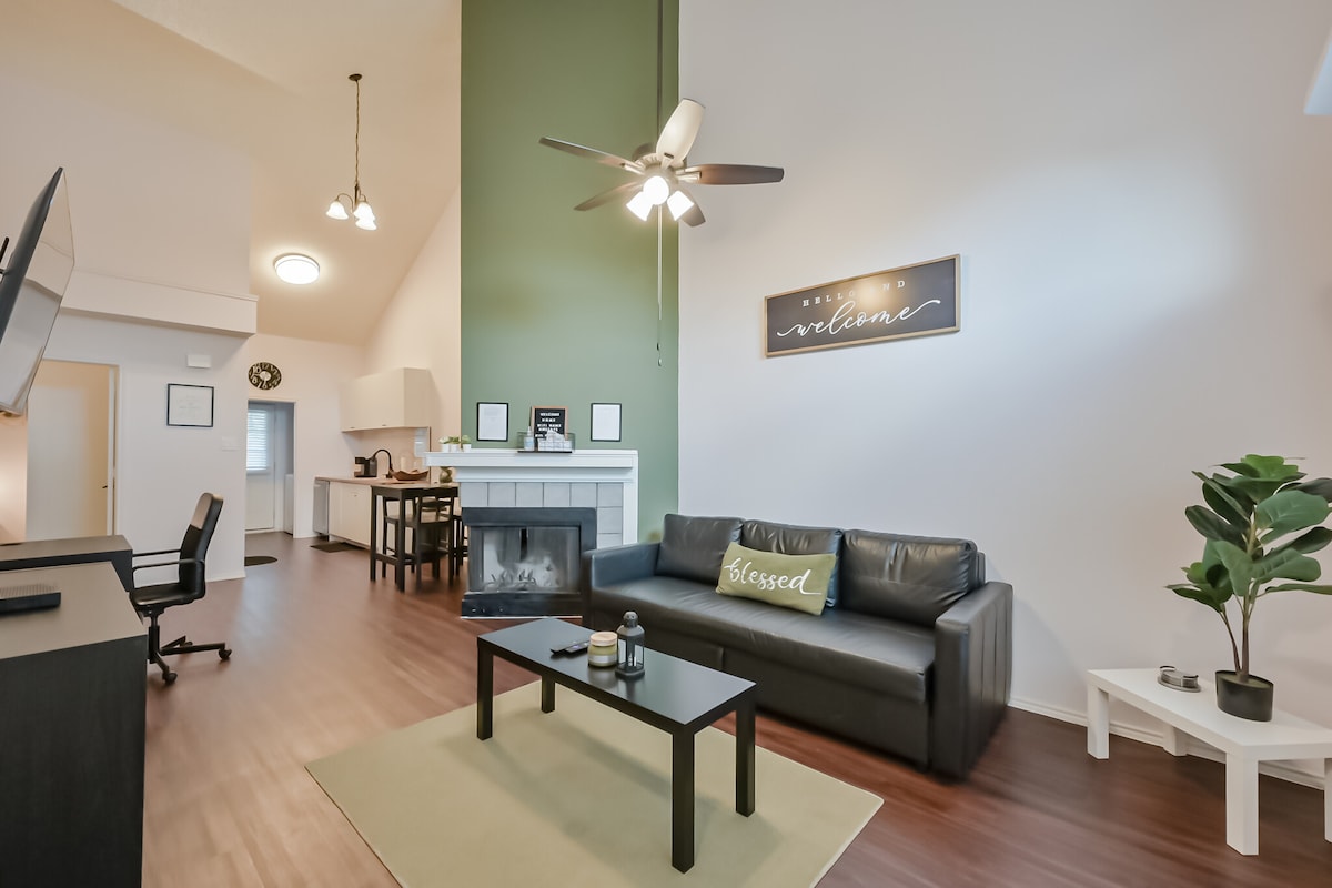 A spacious living area features a comfortable dark sofa with a decorative pillow labeled 'Blessed.' A coffee table sits in front, and a ceiling fan provides ventilation. The room is accented by green walls and a fireplace, creating a cozy atmosphere.