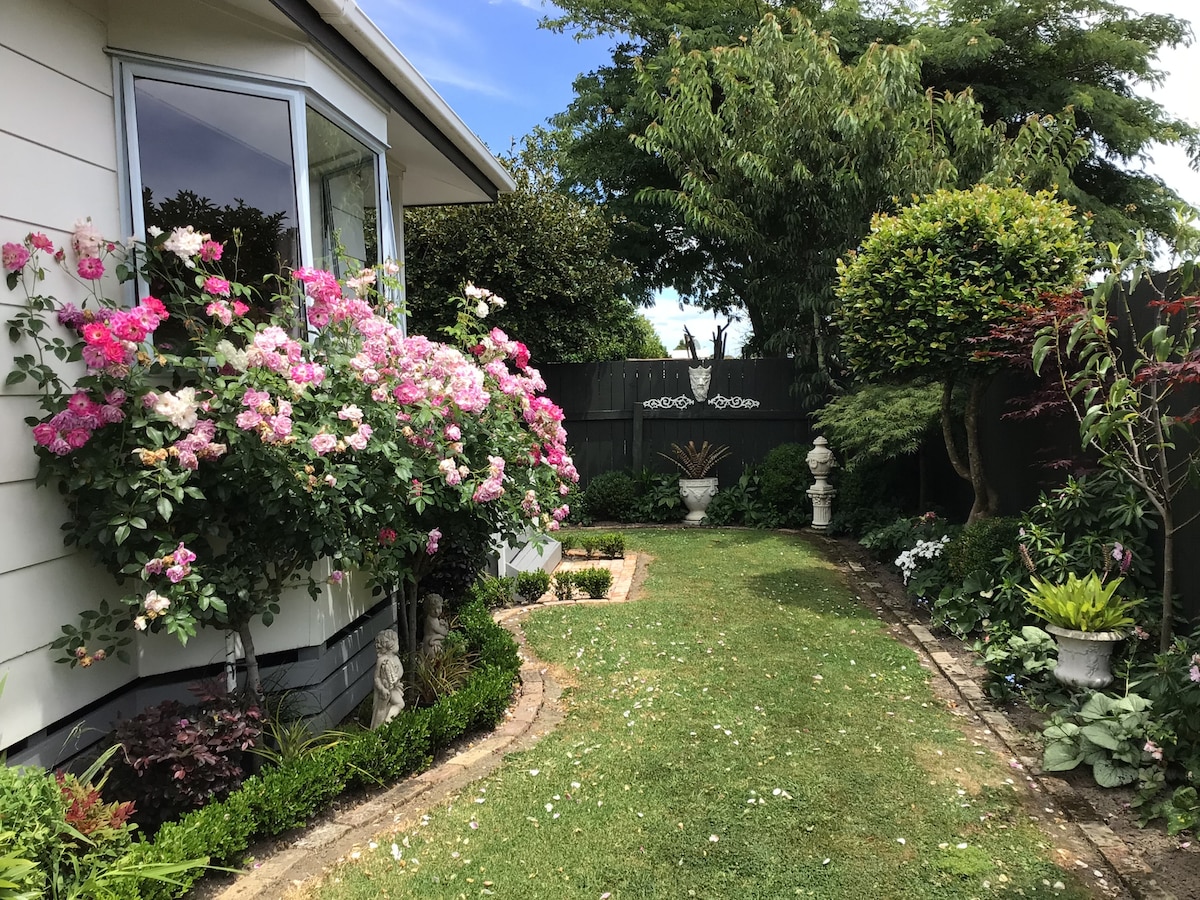 A well-maintained garden is framed by a charming home, with a variety of blooming flowers, particularly pink roses, climbing along the side. Lush greenery surrounds a neatly paved pathway, leading to decorative elements like statues and manicured shrubs, creating a serene outdoor space.