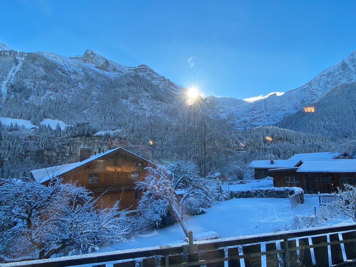 A scenic view showcases snow-covered mountains under a clear blue sky, with sunlight peeking through the peaks. Traditional wooden chalets are surrounded by trees and snow, presenting a serene winter landscape.