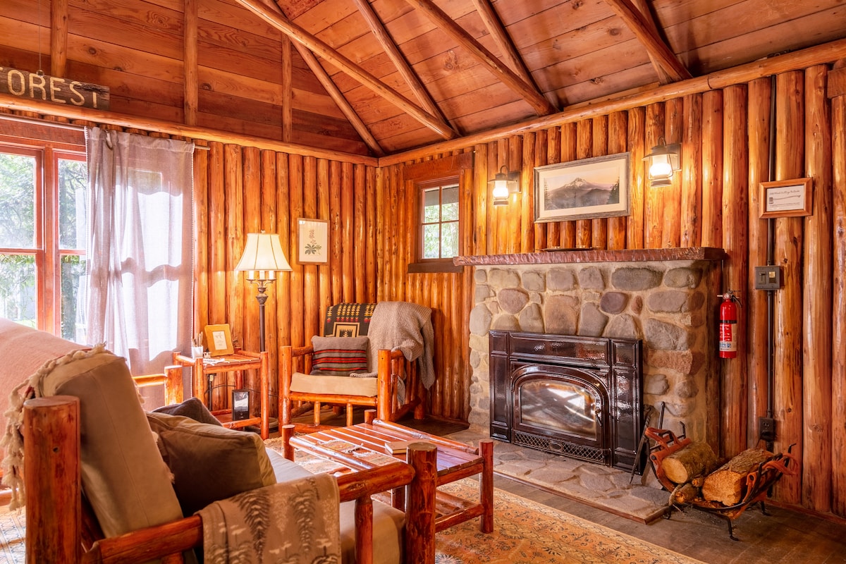 A cozy living area is adorned with warm log walls and a vaulted ceiling featuring crisscross beams. A stone fireplace with a wood-burning stove serves as a central feature. Soft lighting from wall sconces creates a welcoming ambiance around rustic furniture.