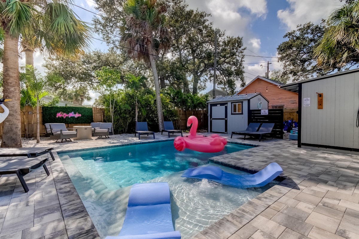 A heated plunge pool is surrounded by chaise loungers and palm trees, offering a relaxing outdoor retreat. A pink inflatable float can be seen resting on the water, while a poolside area includes a shed and shade trees, enhancing the tranquil atmosphere.