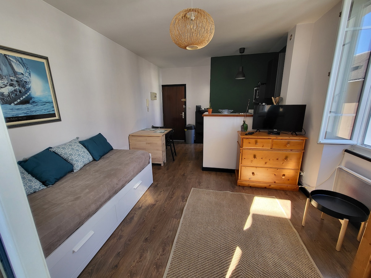 The living area features a light-colored wall complemented by a framed nautical painting. A spacious couch bed is positioned against one wall, paired with decorative cushions. A wooden dresser and a compact table are present, with natural light illuminating the space through an adjacent window.