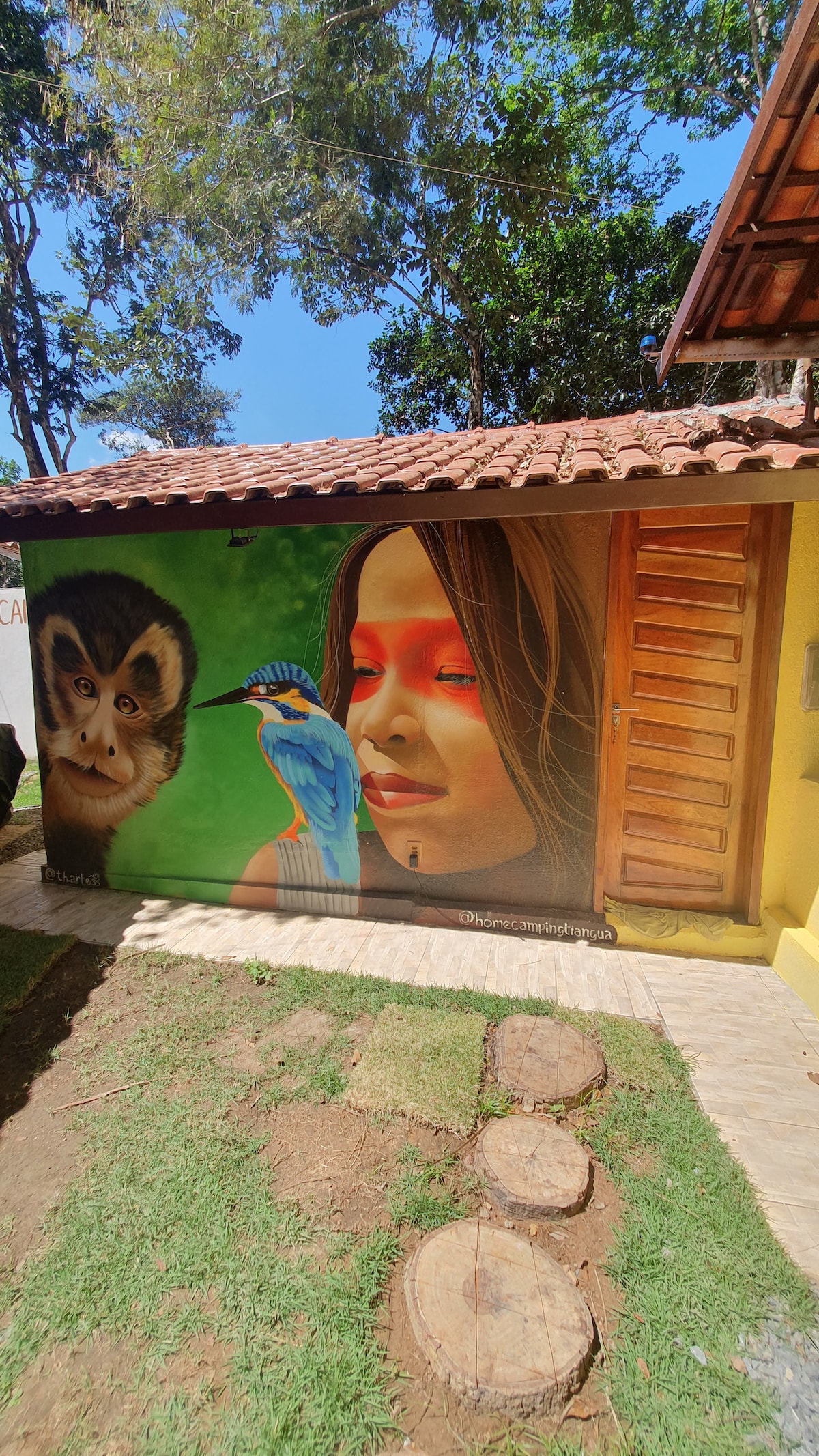 A vibrant mural depicting a bird is painted on an exterior wall, showcasing rich colors against a bright green background. The structure features wooden doors and a tiled roof, with a pathway framed by circular stepping stones on the grassy area.