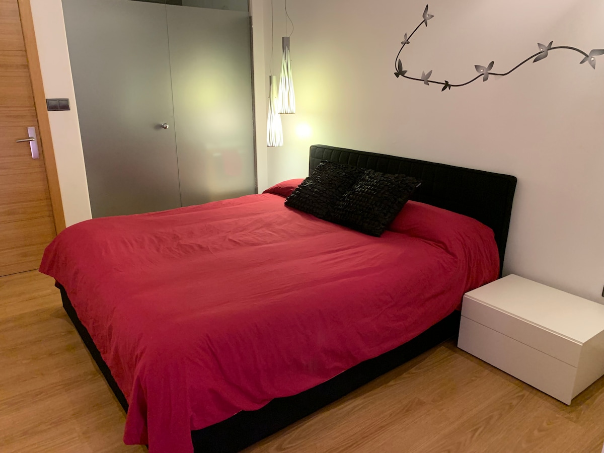 A spacious bed covered with a rich red duvet is centered in the room. Soft lighting from a pendant fixture and bedside table illuminates the space. A modern side table stands beside the bed, complementing the overall decor.