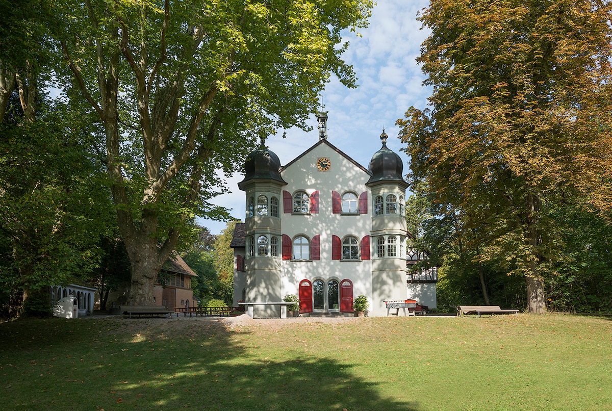 An elegant two-story building features distinctive onion-shaped domes and bright red shutters. The structure is complemented by lush greenery, with tall trees framing the scene and a grassy area in the foreground. Natural light highlights the architecture against a partly cloudy sky.