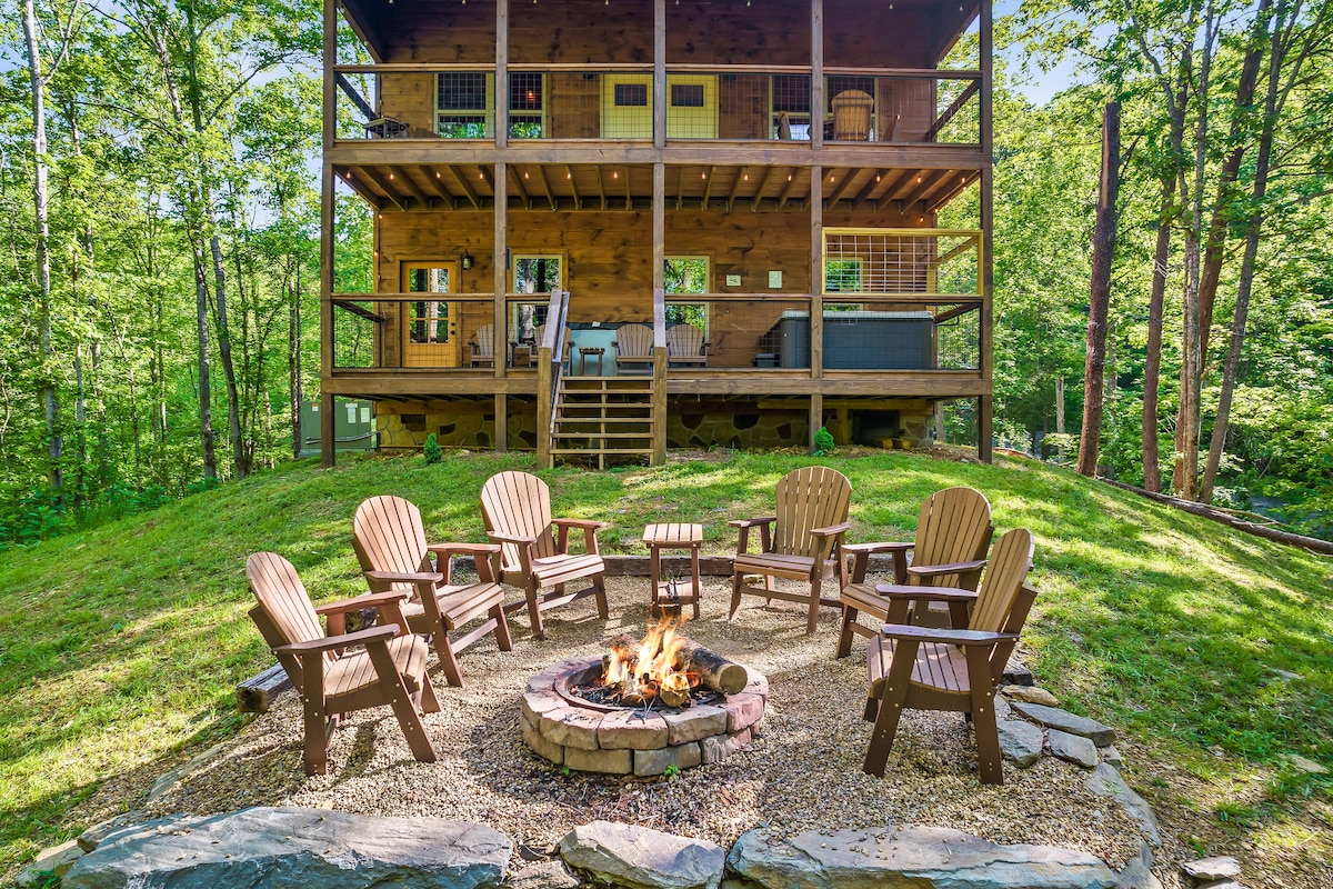 A backyard fire pit area is arranged with sturdy wooden chairs positioned in a circular formation. A stone border encircles the fire pit, which rests on a gravel surface. The cabin features three covered decks above, surrounded by lush greenery.