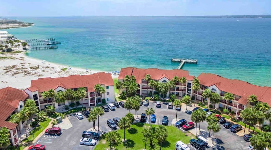 An aerial view captures the condo community with red-tiled roofs set against the backdrop of the Santa Rosa Sound. Lush palm trees surround the property, while a private pier extends into the water. The sandy beach is visible in the distance, highlighting the tranquil coastal setting.