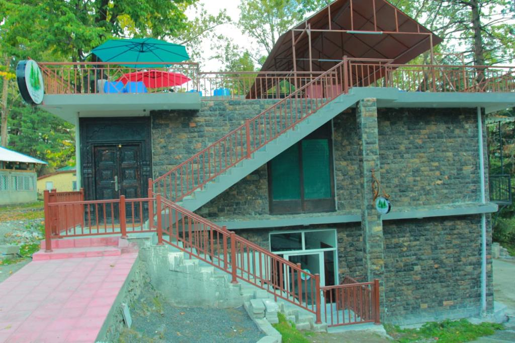 A two-story building is presented, featuring a stone facade. A set of red metal stairs leads to a rooftop terrace with colorful umbrellas. The entrance is accentuated by a dark wooden door, framed by large glass windows on the ground floor.