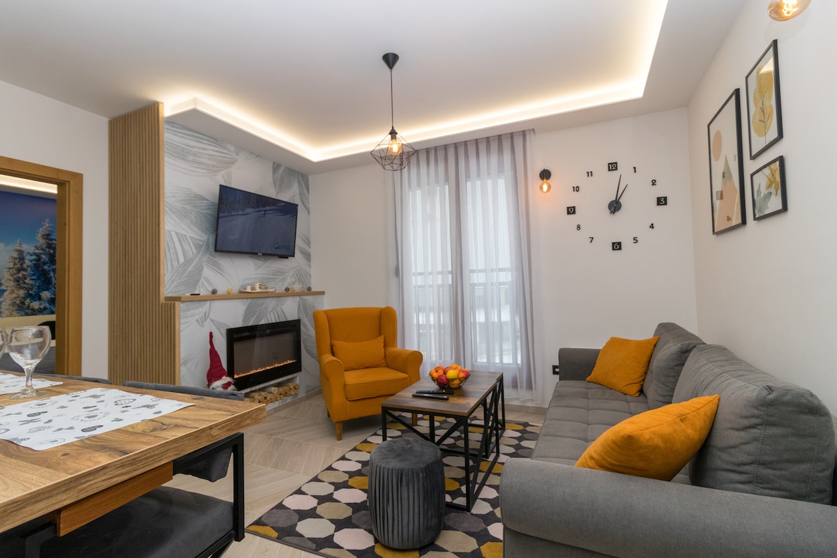 A living area is presented with a comfortable gray sofa, bright orange armchair, and a patterned rug. A modern fireplace serves as a focal point, while a wall-mounted television is visible above. Natural light filters through sheer curtains, enhancing the welcoming ambiance.