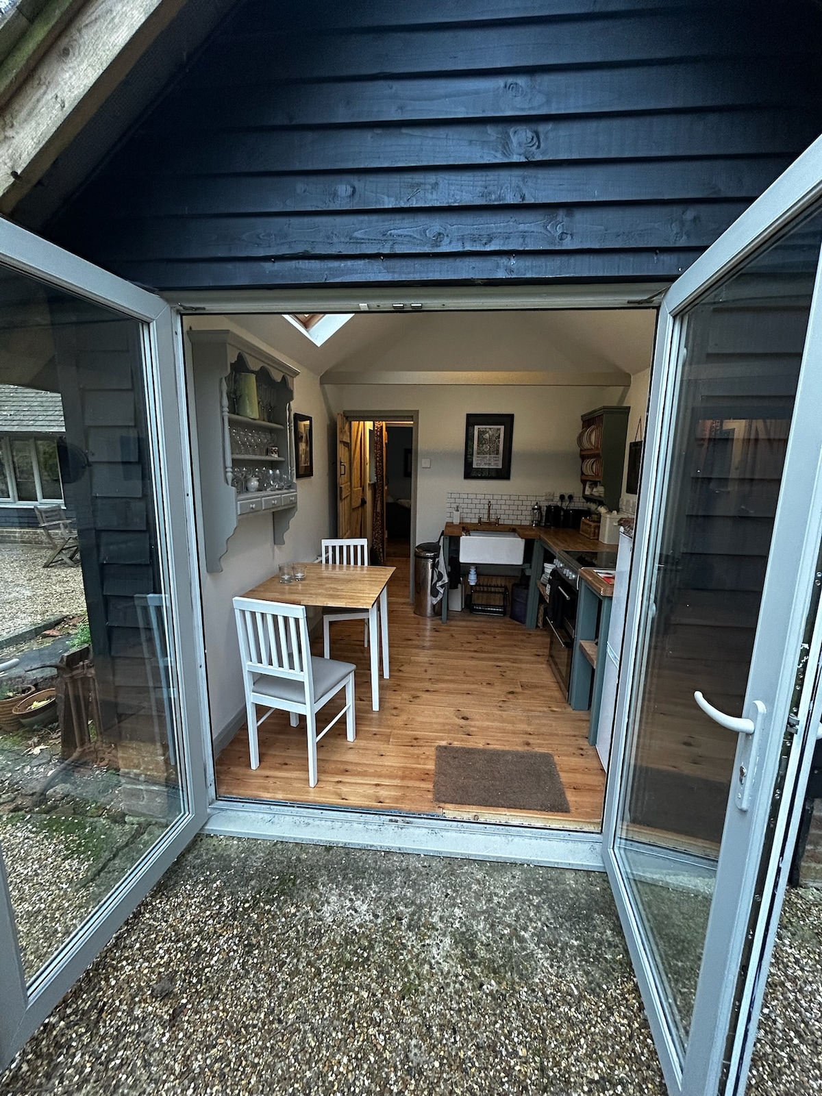 A view into the kitchen showcases a wooden dining table with four chairs and an inviting atmosphere. Natural light fills the space through a skylight, illuminating the wood flooring. Various kitchen appliances and storage units are organized along the walls.