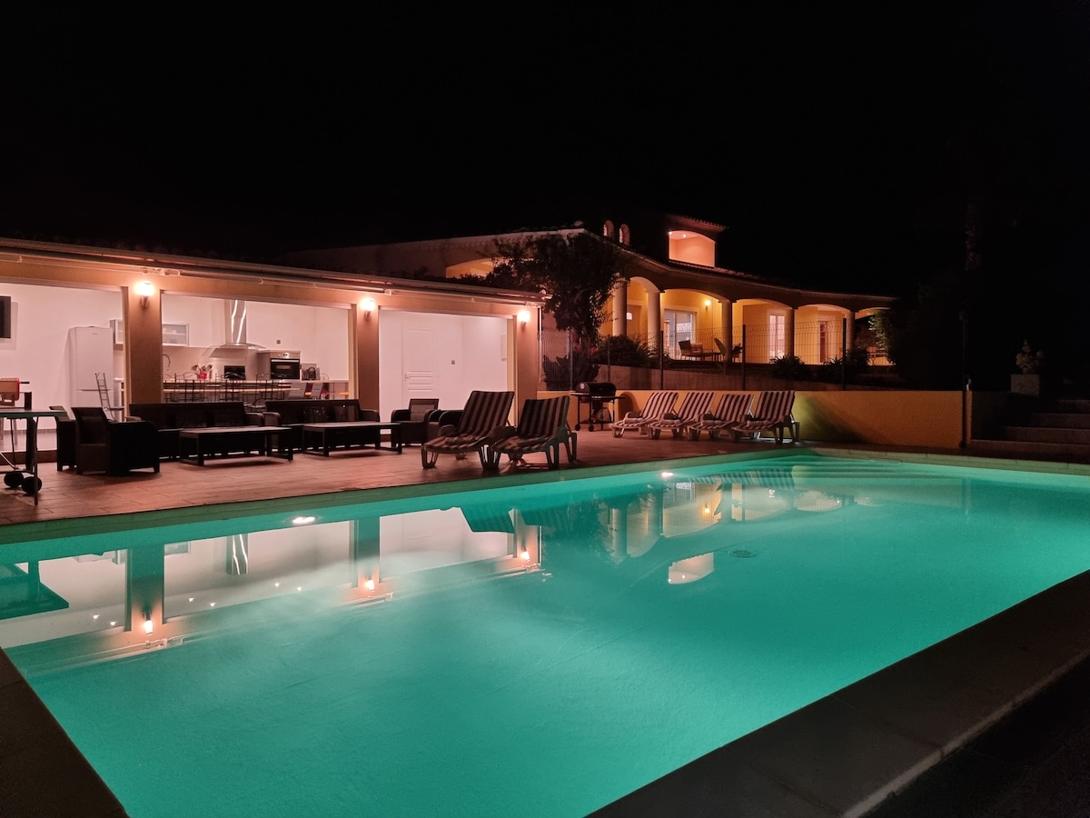 A private pool is illuminated in turquoise, reflecting the warm lights of a spacious villa. The outdoor area is furnished with lounge chairs and a dining table, providing an inviting setting. The villa features large windows, showcasing the interior spaces.