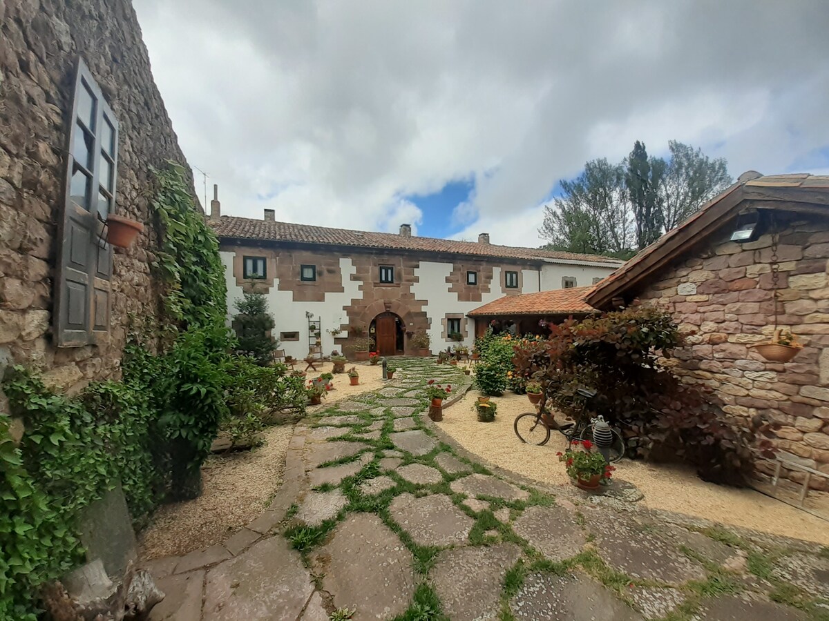 A charming courtyard features a winding stone pathway surrounded by greenery and flower pots. Two rustic stone buildings frame the entrance, with a welcoming archway visible in the background. The serene atmosphere is enhanced by soft clouds overhead, providing a peaceful backdrop.