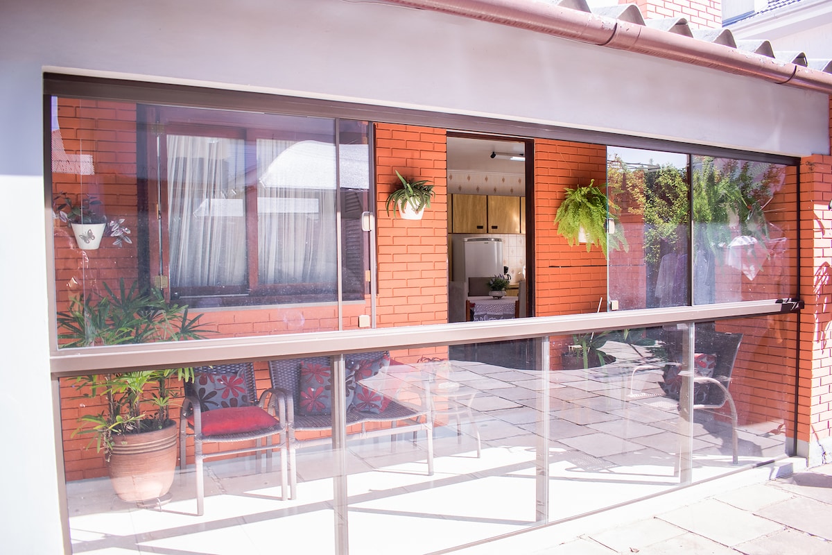 A transparent glass enclosure is showcased, revealing a patio area with two chairs adorned with colorful cushions. Potted plants hang from the exterior wall, complementing the brick structure and inviting natural light into the space.