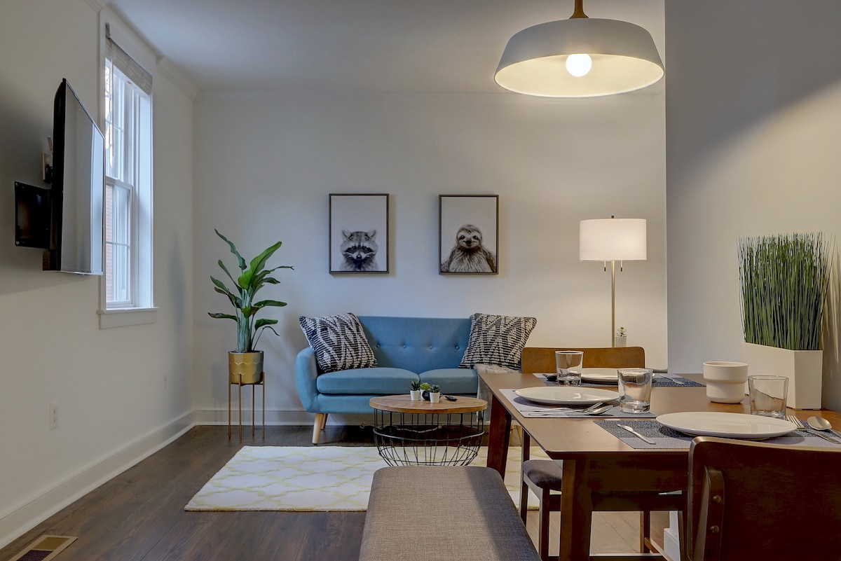A welcoming living and dining area features a blue sofa with decorative pillows and two framed animal prints above it. A wooden dining table is set for four, accompanied by a neutral-toned runner. A green plant and a lamp add warmth to the space.