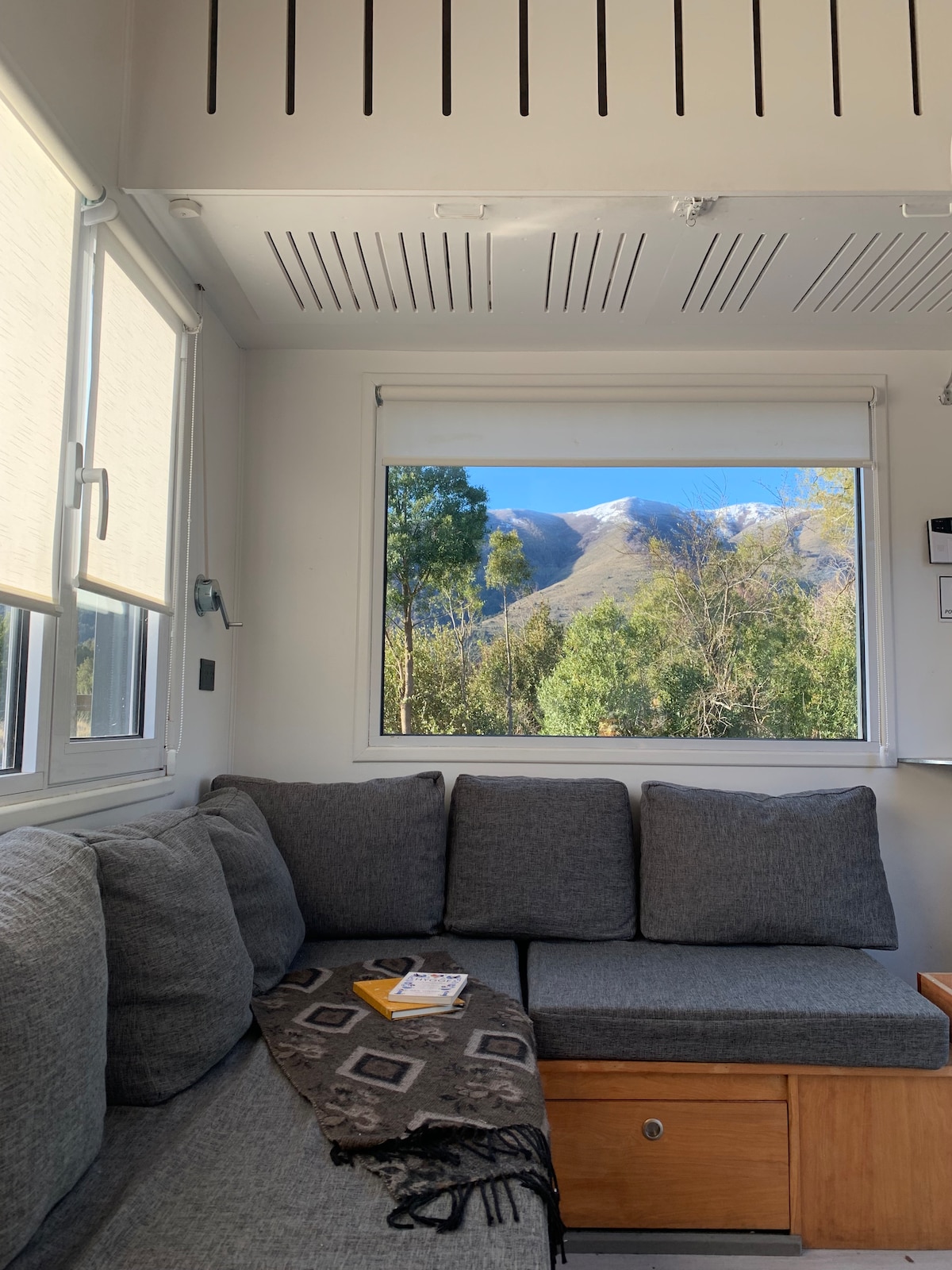 A cozy living area features a grey sectional sofa, complemented by a warm throw blanket. A large window reveals a scenic view of snow-capped mountains and lush greenery, enhancing the natural light in the space.