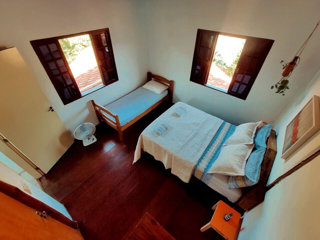 A well-lit sleeping area features a spacious bed and a single bed, both made with white linens. Natural light enters through two windows, framed in dark wood. A small bedside table with a decorative item is positioned beside the larger bed, enhancing the room's tidy appearance.