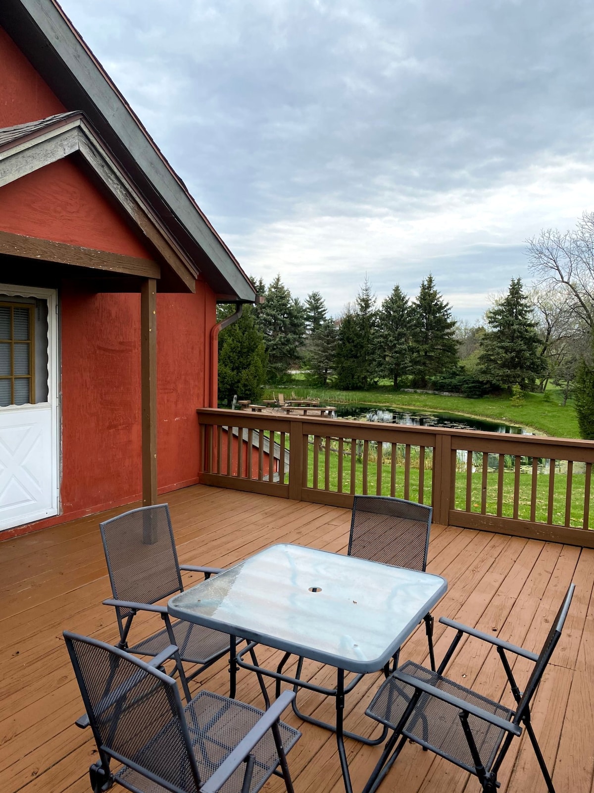 A spacious deck features a glass-top table surrounded by four metal chairs. The peaceful pond and lush greenery are visible beyond the railing, complemented by a cloudy sky overhead.