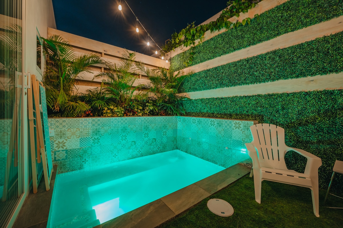 A private oasis terrace is showcased, featuring a plunge pool illuminated in soft turquoise light. Lush greenery surrounds the area, complemented by outdoor string lights overhead. A comfortable white chair is positioned nearby, enhancing the relaxing ambiance of this secluded space.