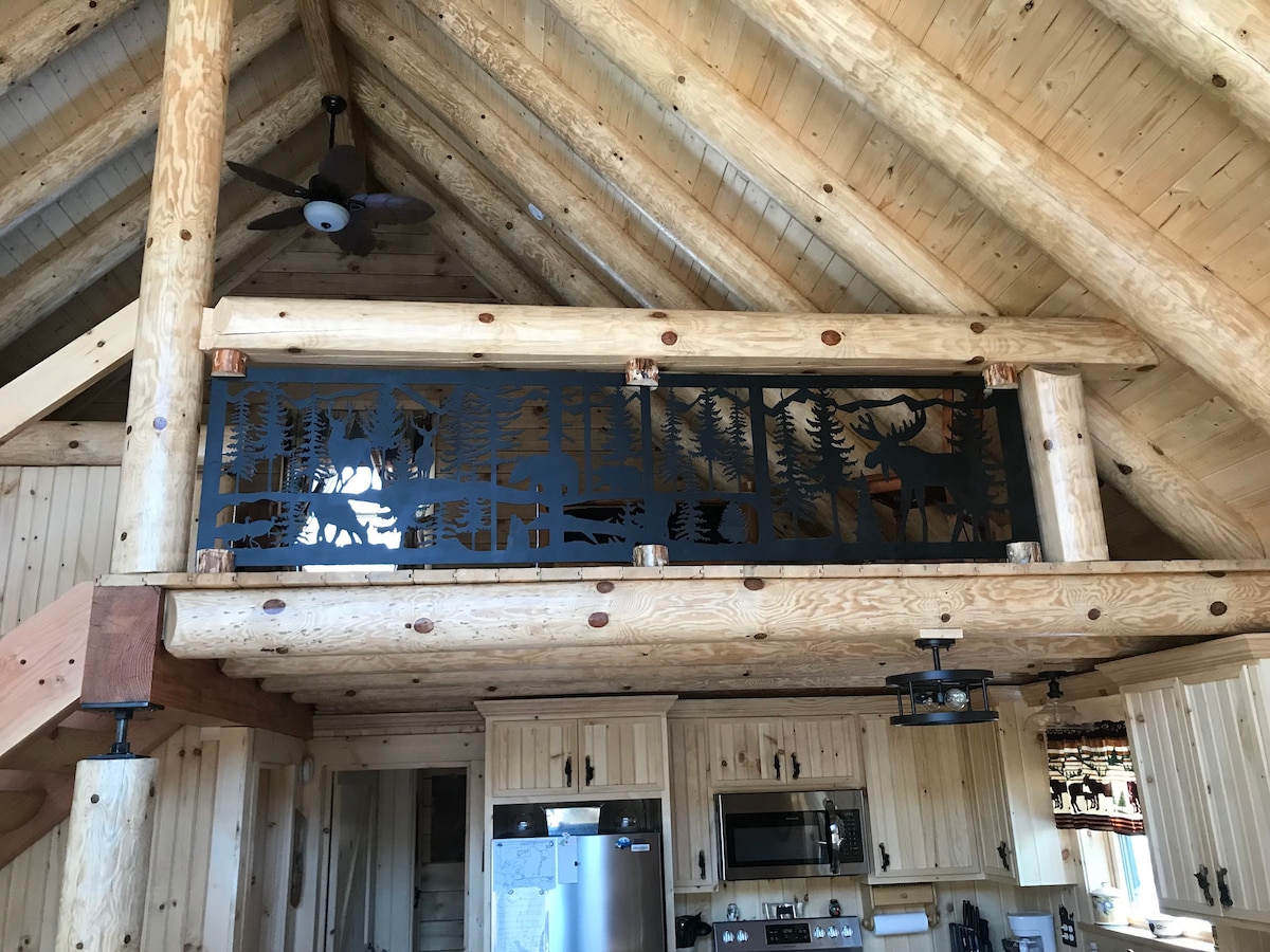 A spacious cabin interior showcases a high, vaulted ceiling with exposed wooden beams. A decorative metal railing, featuring nature-inspired designs, overlooks the lower living area. Light wood cabinetry and appliances are visible, contributing to a warm and rustic ambiance.