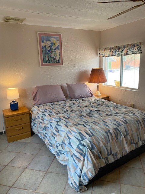 A cozy bedroom features a Queen-sized bed with a patterned coverlet. Two nightstands flank the bed, each with a lamp providing soft light. Natural light enters through a nearby window adorned with floral curtains, adding warmth to the space.