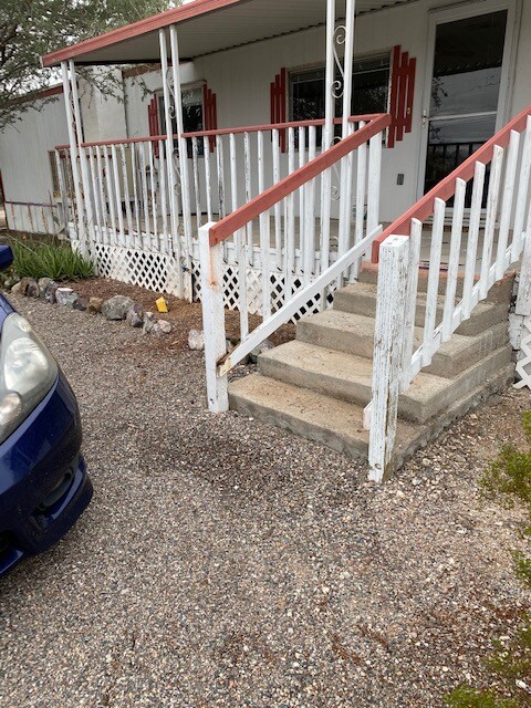 A front porch area is shown with a set of steps leading up to the entrance of the property. The porch is bordered by white railings with red accents. A gravel pathway runs alongside, providing access for parking and walking.