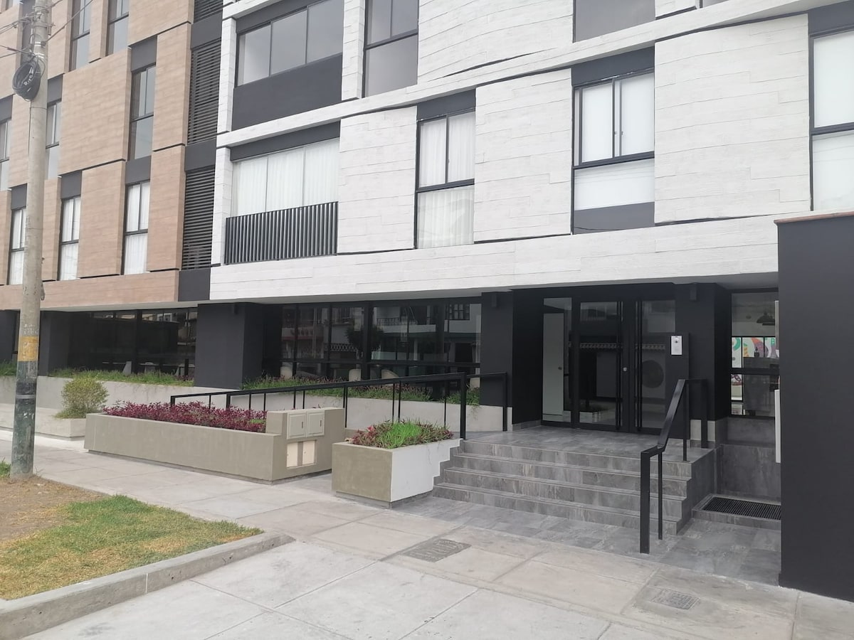 The building's exterior is characterized by a modern architectural design, featuring large windows and a combination of light and dark materials. A wide staircase leads to the entrance, framed by landscaped planters filled with greenery and colorful flowers, creating an inviting approach.