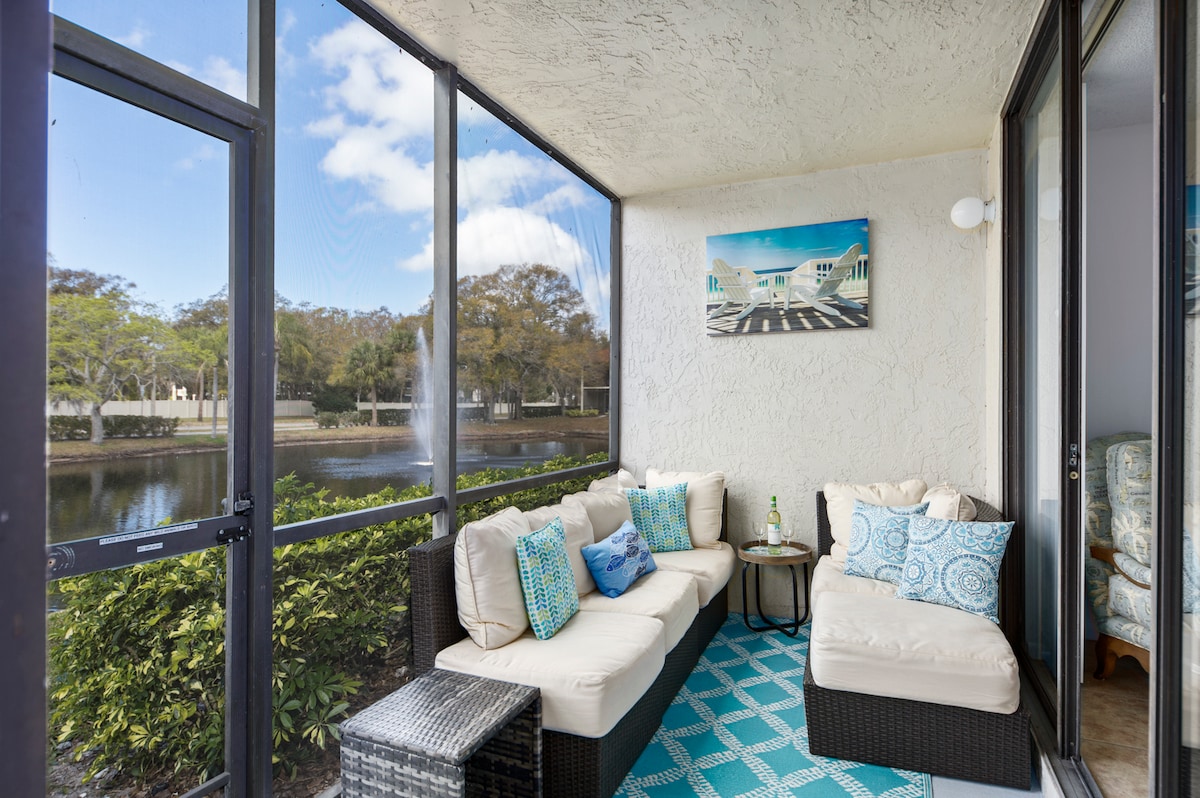 A screened-in porch features a lounging area with light-colored cushions and decorative blue pillows. A glass-top table rests on a patterned mat. Views of a serene lake and lush greenery are visible through the screens, allowing for a bright and airy atmosphere.